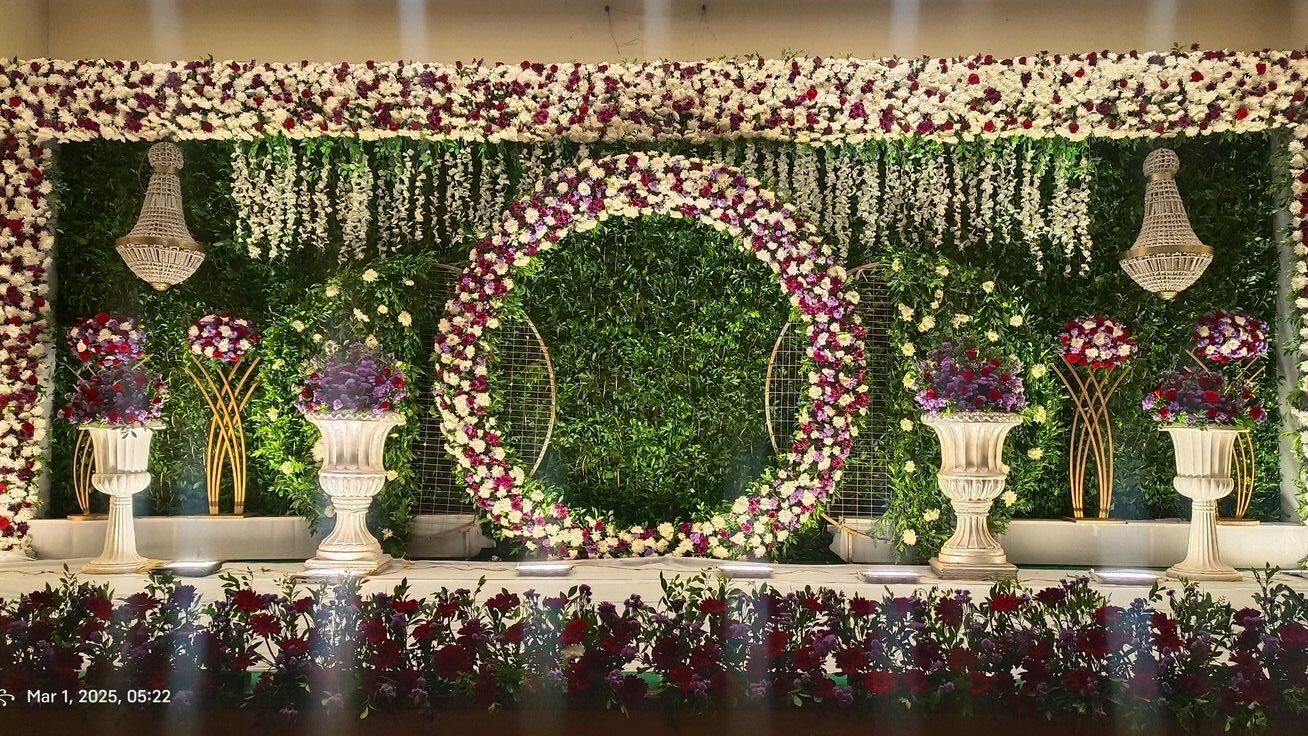 Elegant floral stage decoration with green wall backdrop, round flower ring, chandeliers, and luxury floral arrangements in Hyderabad.